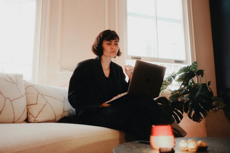 Woman in suit using laptop on sofa in bright room, modern lifestyle.