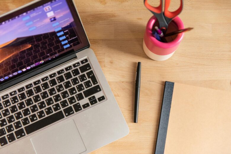 Top-down view of a laptop, pen, notebook, and scissors on a wooden desk, depicting a workspace setup.
