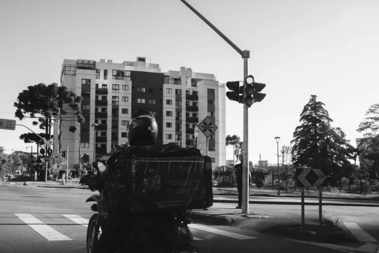 Pessoa entregadora de aplicativo em moto, com bolsa térmica presa na garupa, parada em via urbana movimentada durante o horário de pico